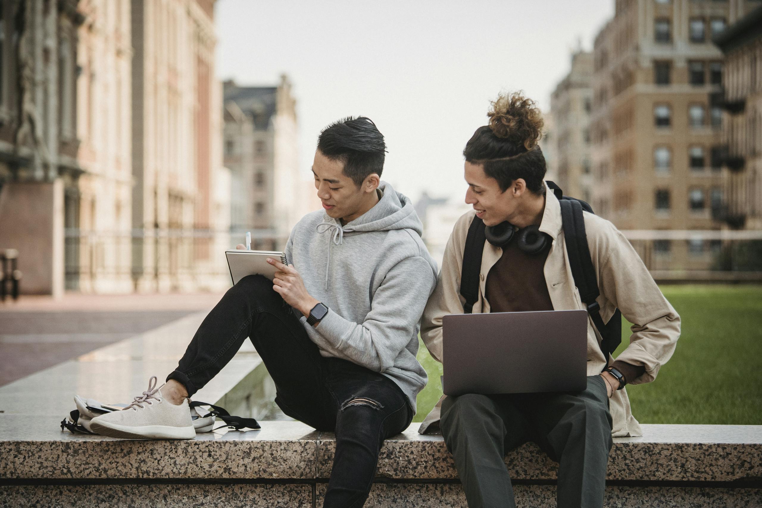 Two young men studying together outdoors with a laptop and notebook in an urban park setting.